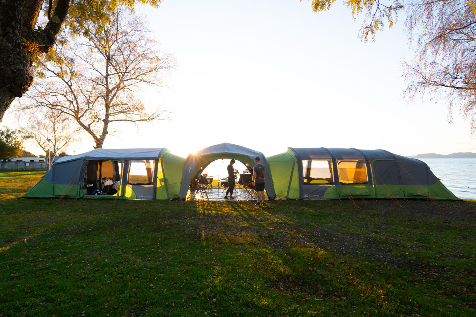 From left to right Tui 4 Air Tent, Savanna 3.5 Air Shelter, and Tui 6 Air Tent. Pitched lakeside on a grass campsite, framed by trees with the sun setting in the background